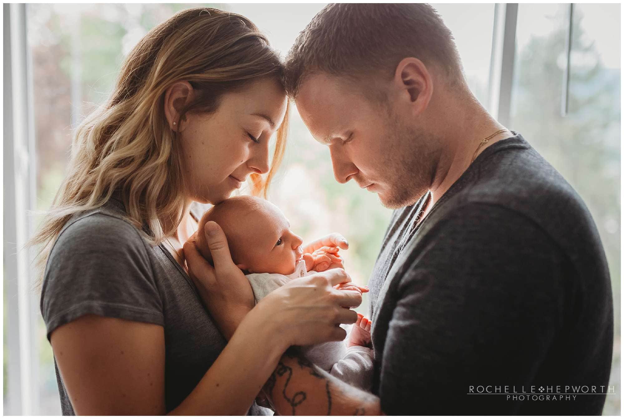 couple standing in front of window with newborn baby between them