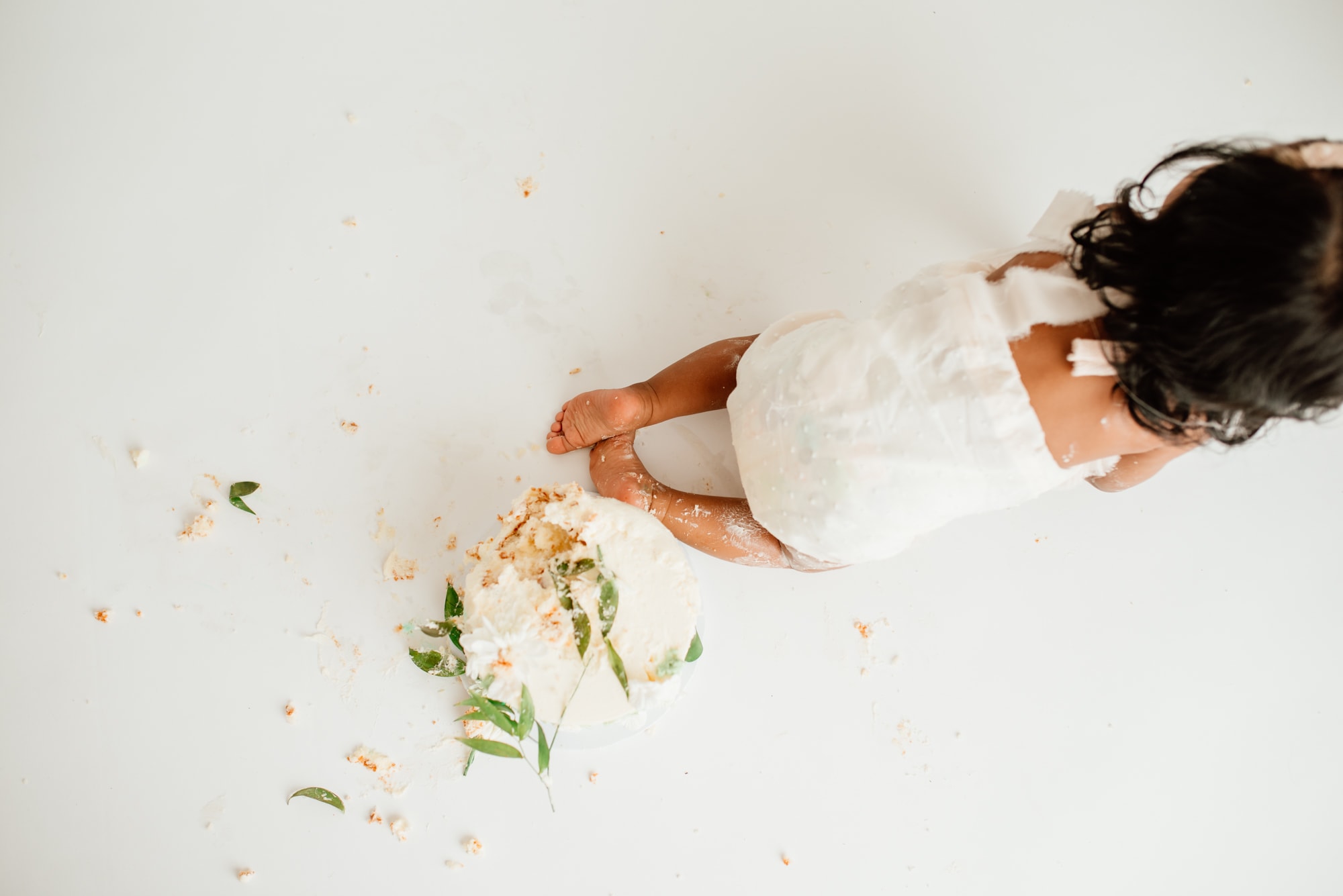 Baby in white romper crawls away from smashed cake in a white studio.