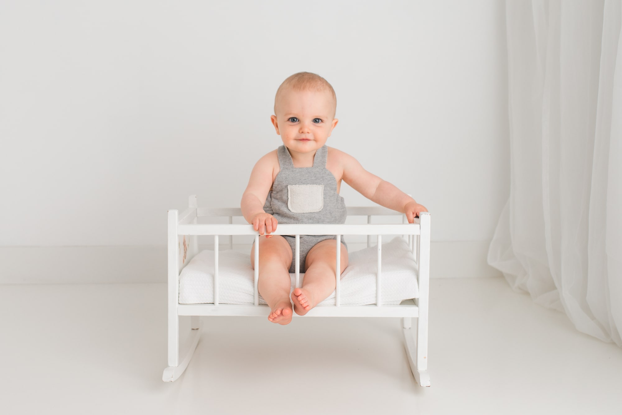 Little boy in grey romper sits in an antique small doll cradle.