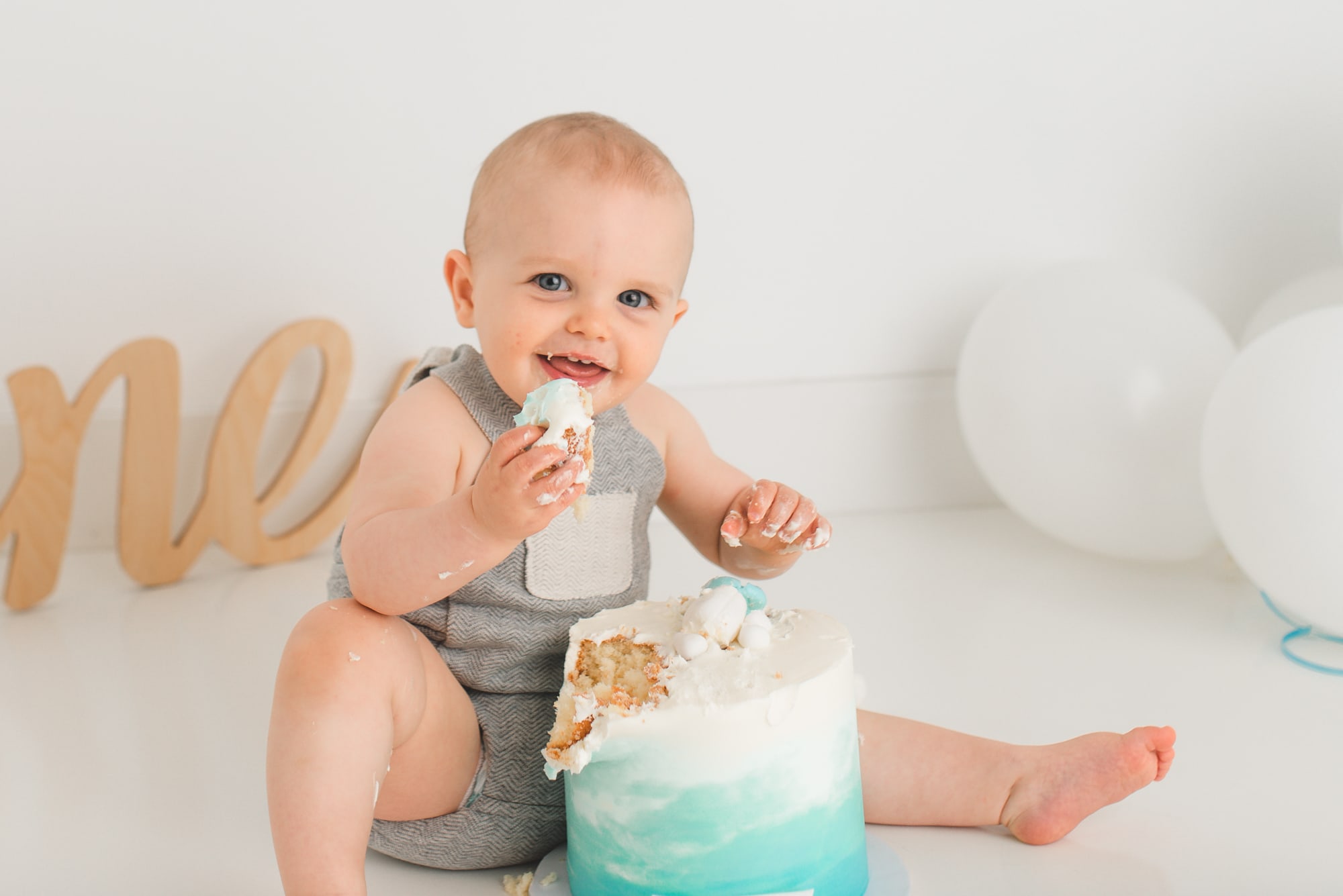 One year old toddler puts big piece of blue and white ombre decorated cake into his mouth while smiling.