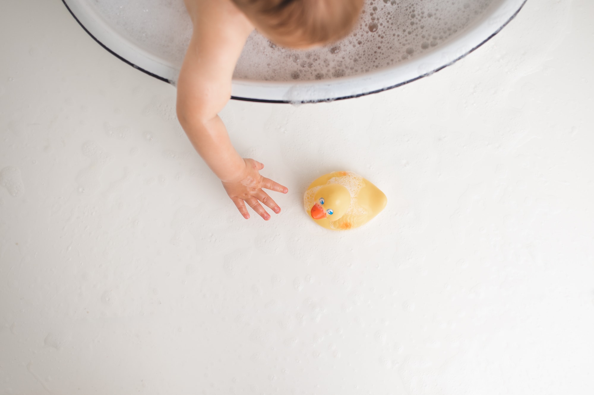Overhead view of a baby's arm reaching out of a bubble bath towards a yellow toy duck.