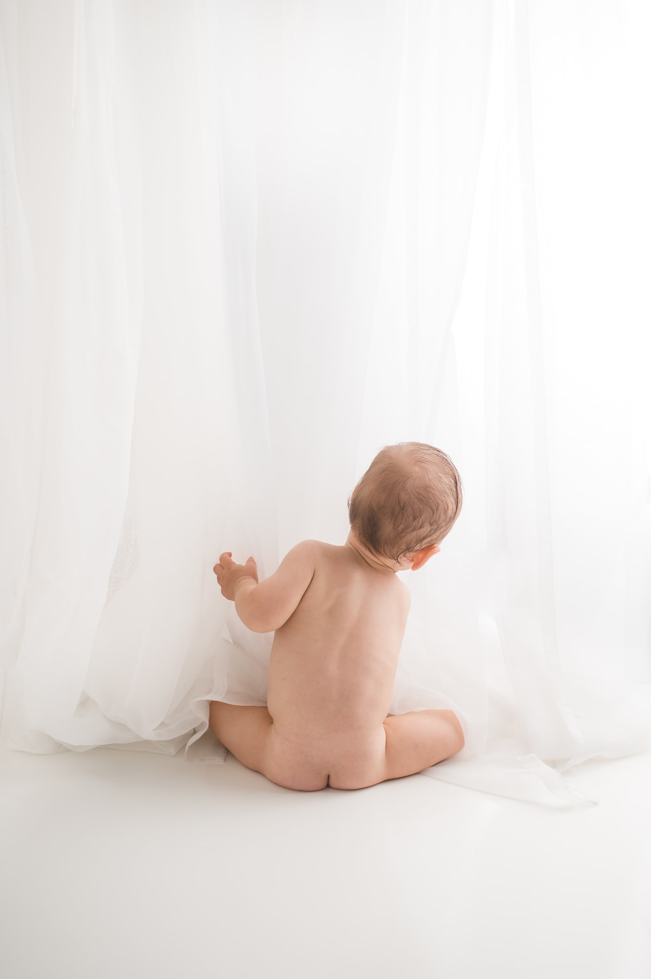 Cute naked baby peeking through white curtains.