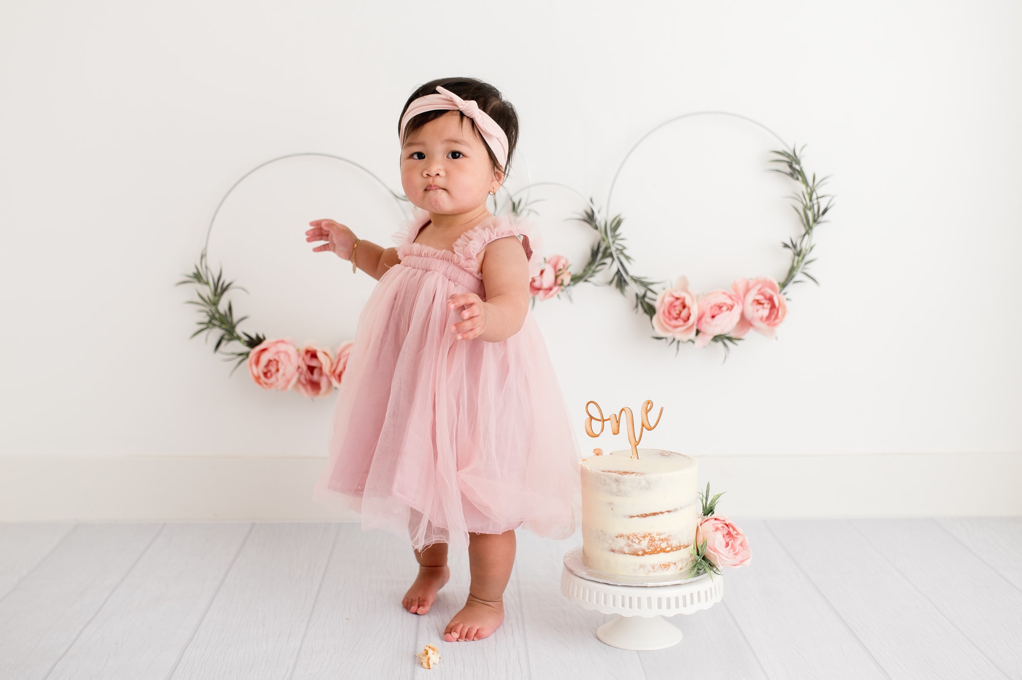 Toddler girl in pink dress stand next to cake and floral hoops at cake smash session.
