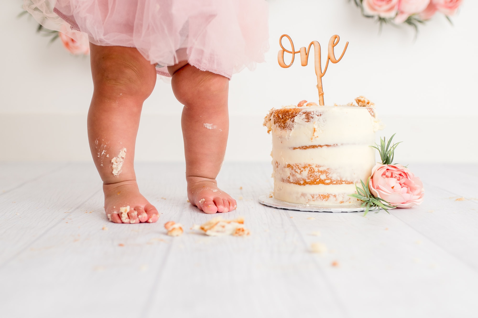 Chubby legs of girl next to a cake that has been smashed and has a cake topper saying "one".
