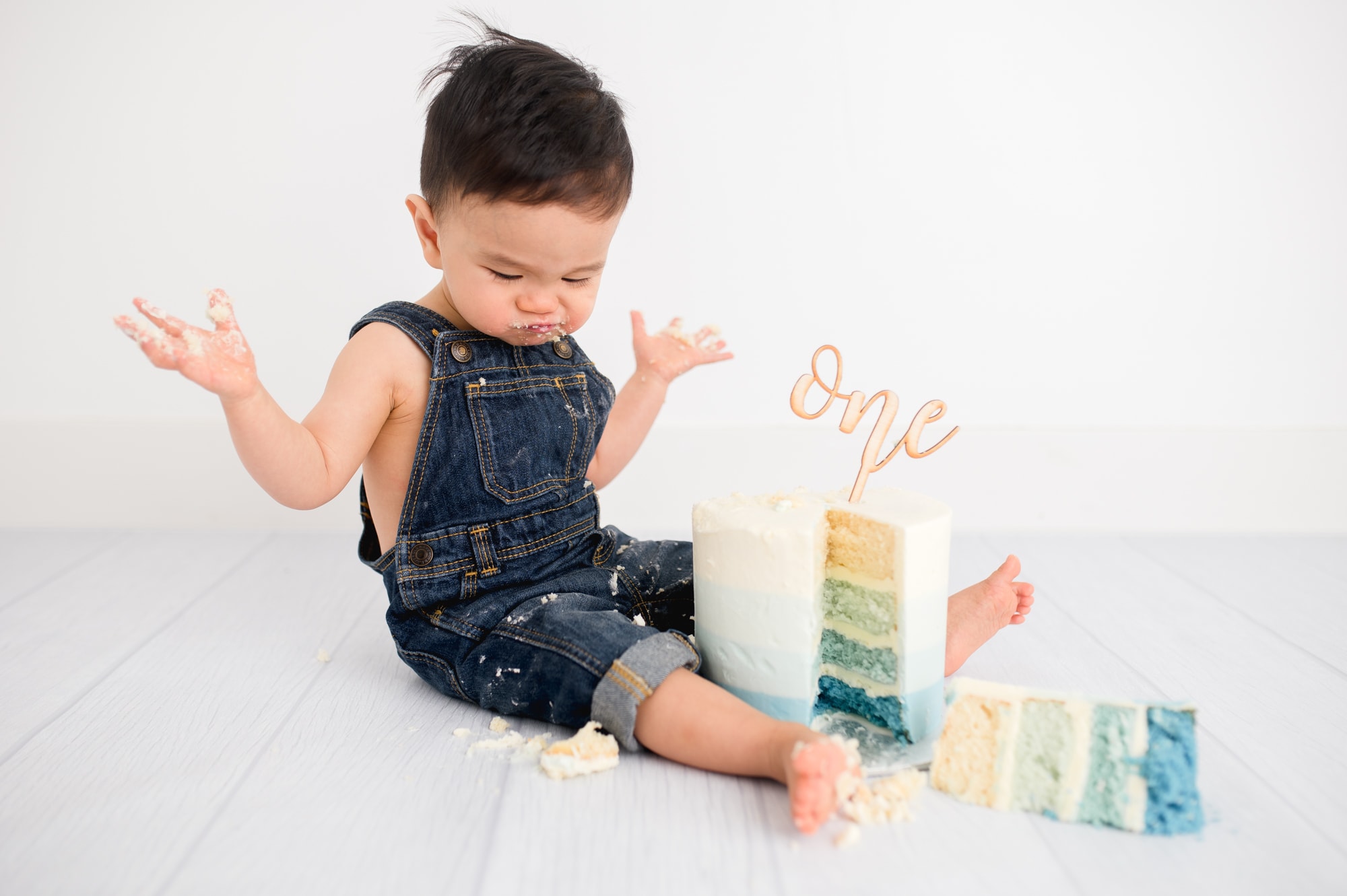 Toddler boy in denim overalls throws his hands in the air after a piece of cake falls in this Vancouver studio session.