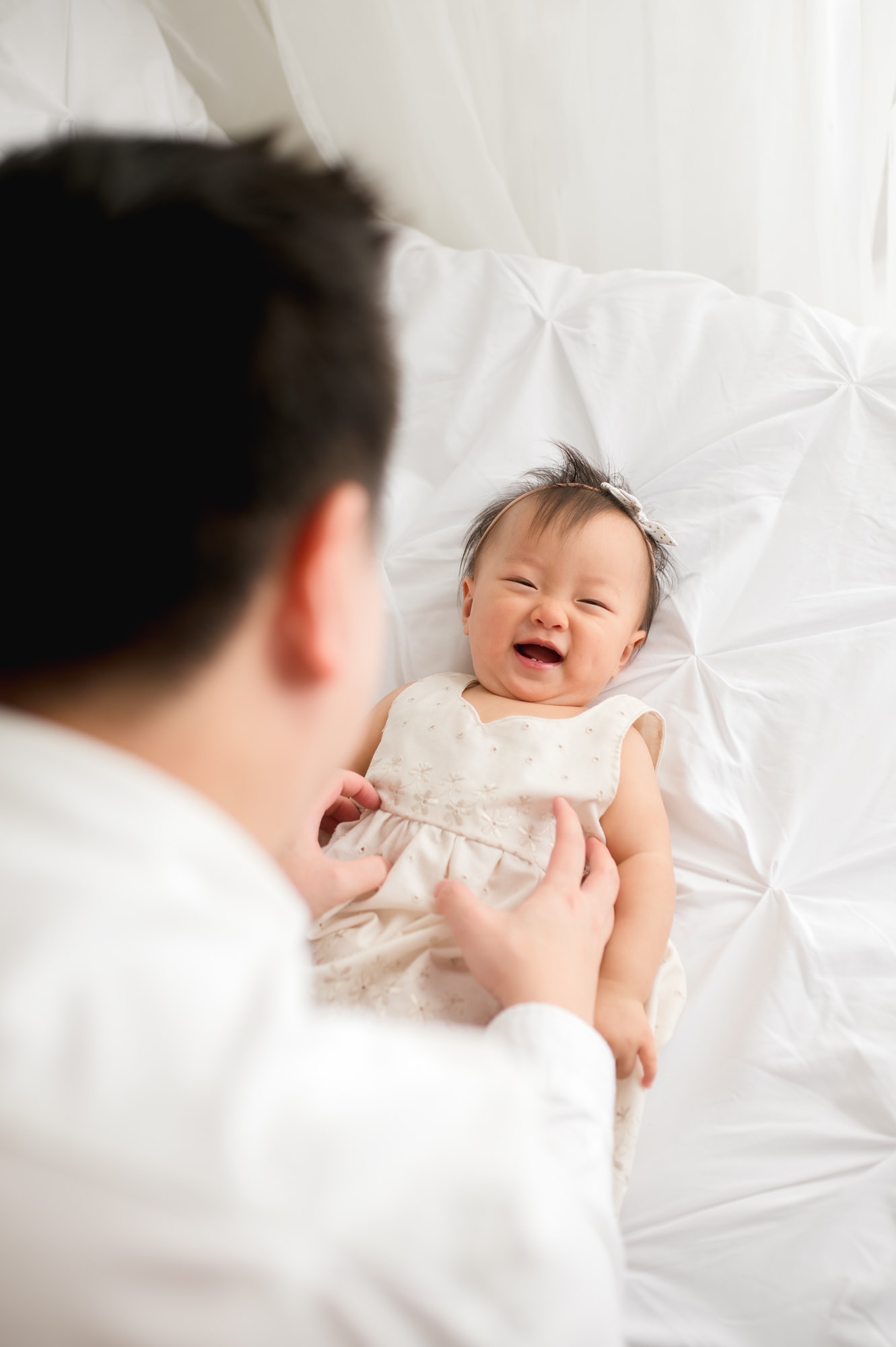 Parent tickles laughing child on bed at their all-white first birthday studio session.