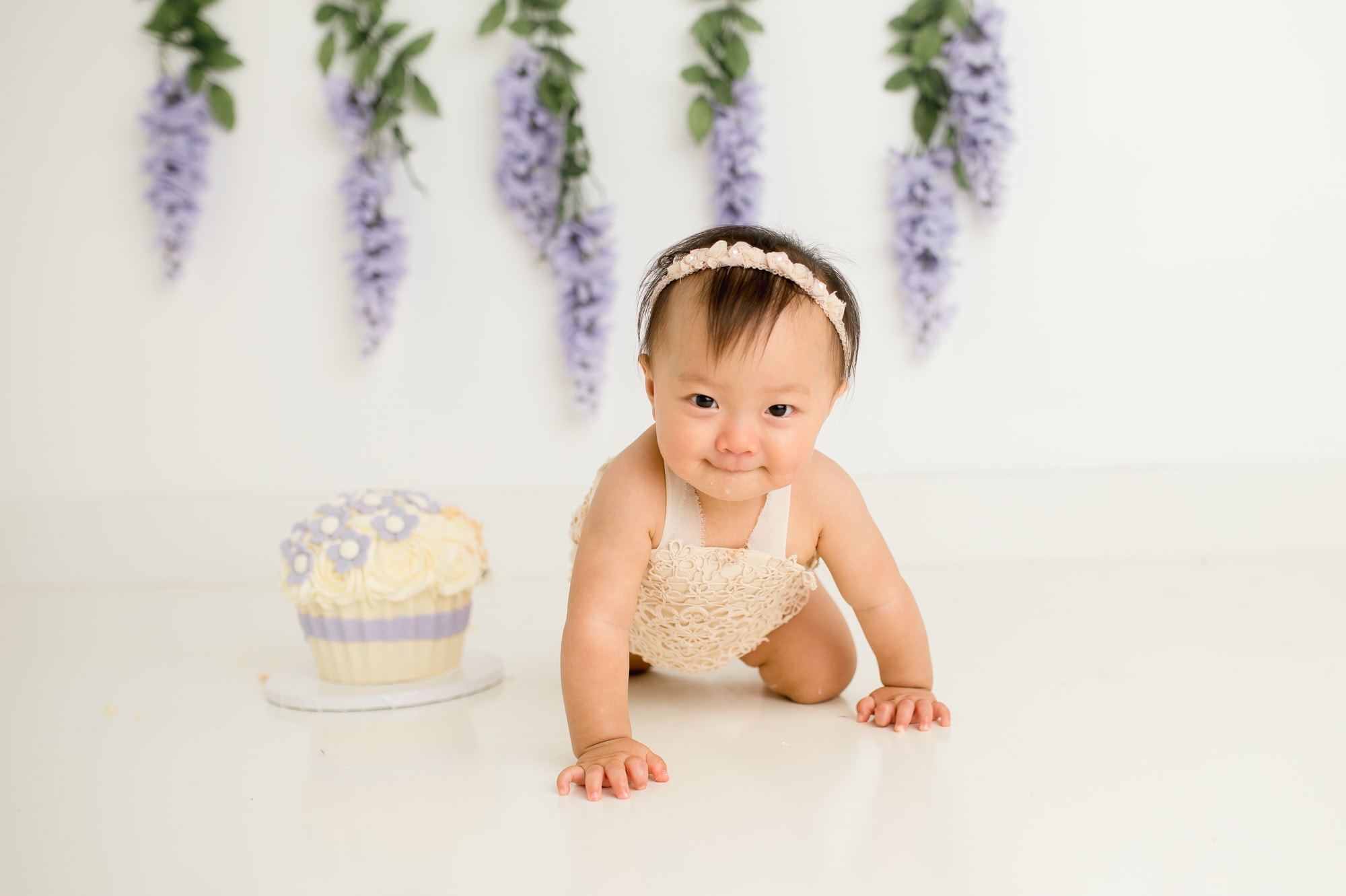 Baby girl crawls towards camera next to a lavender and white cake and hanging purple wisteria backdrop.