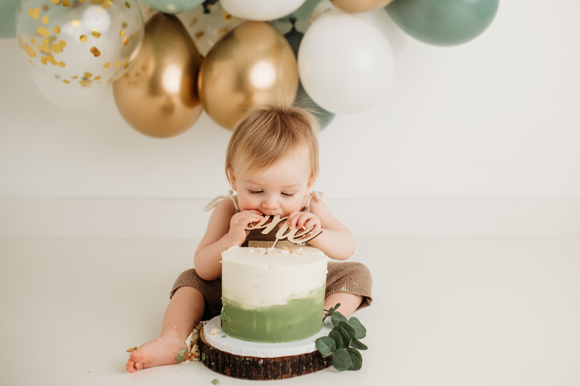 Boy eats whole cake in front of a green, gold and white balloon garland.