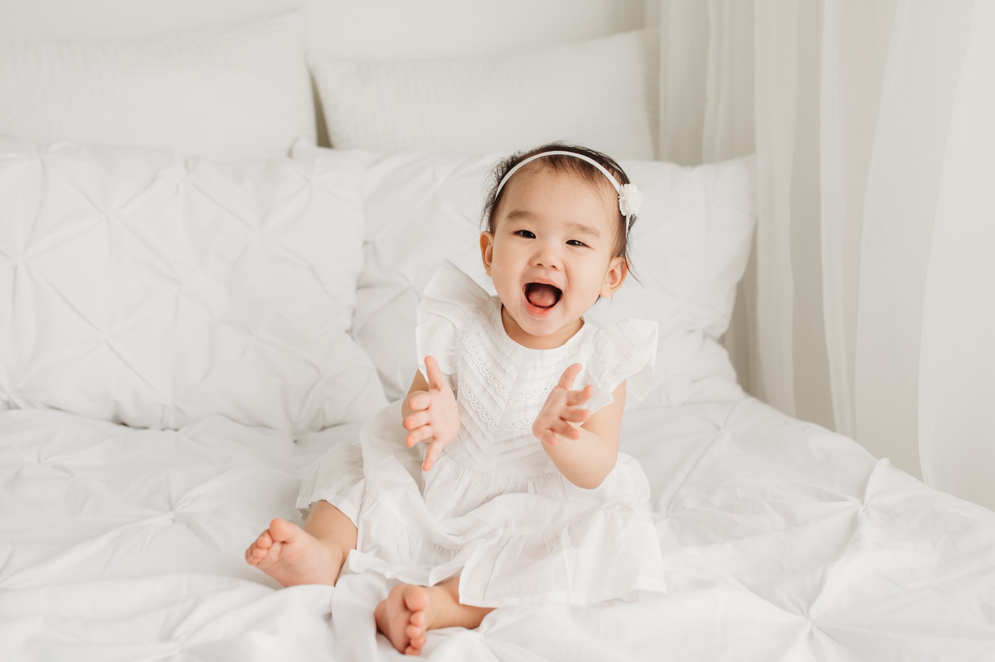 Asian toddler girl laughs happily on a white bed in this Port Coquitlam portrait session.