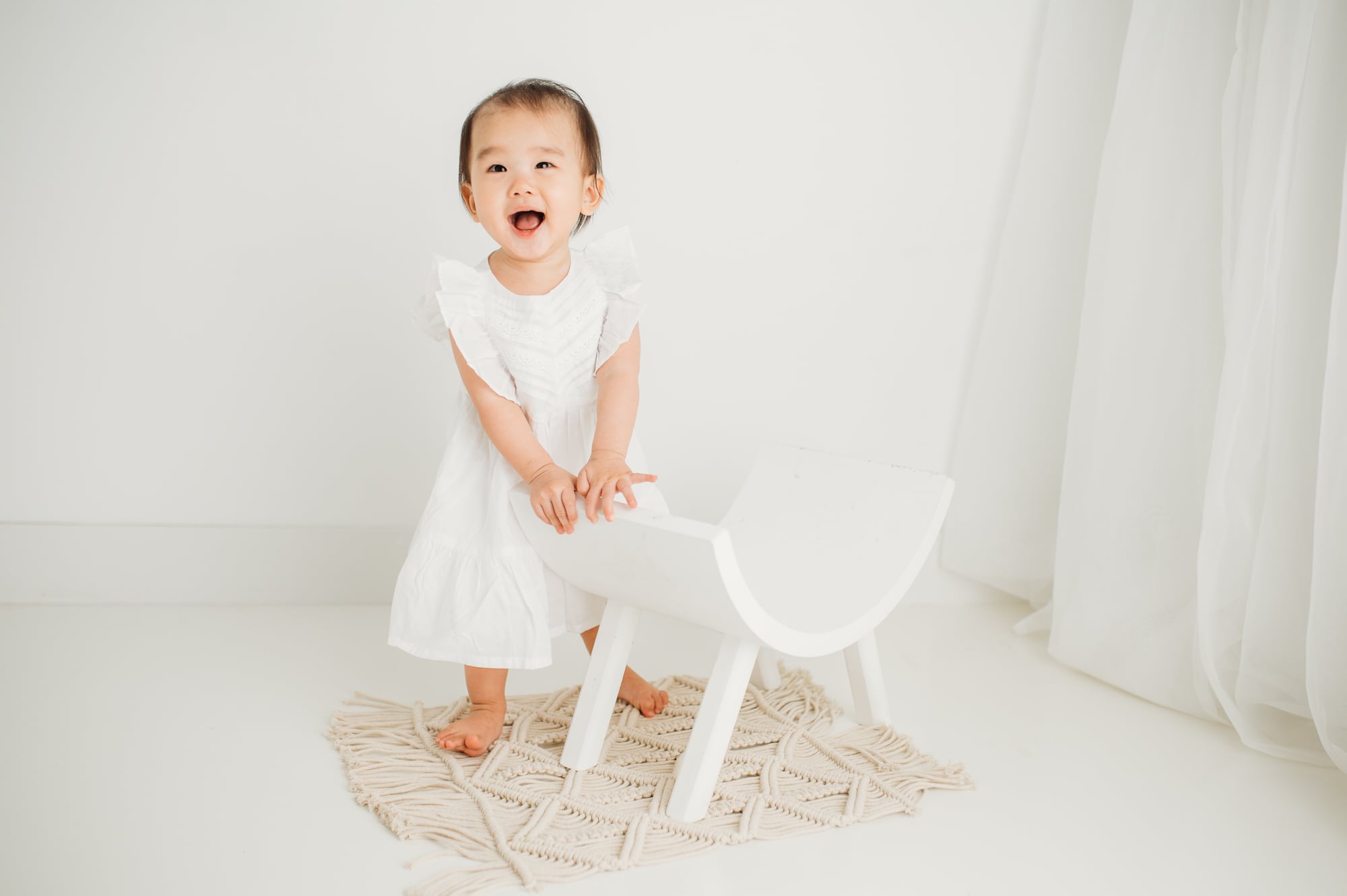 Asian girl laughs in a milestone portrait taken for her first birthday session, standing next to a white bendy chair.