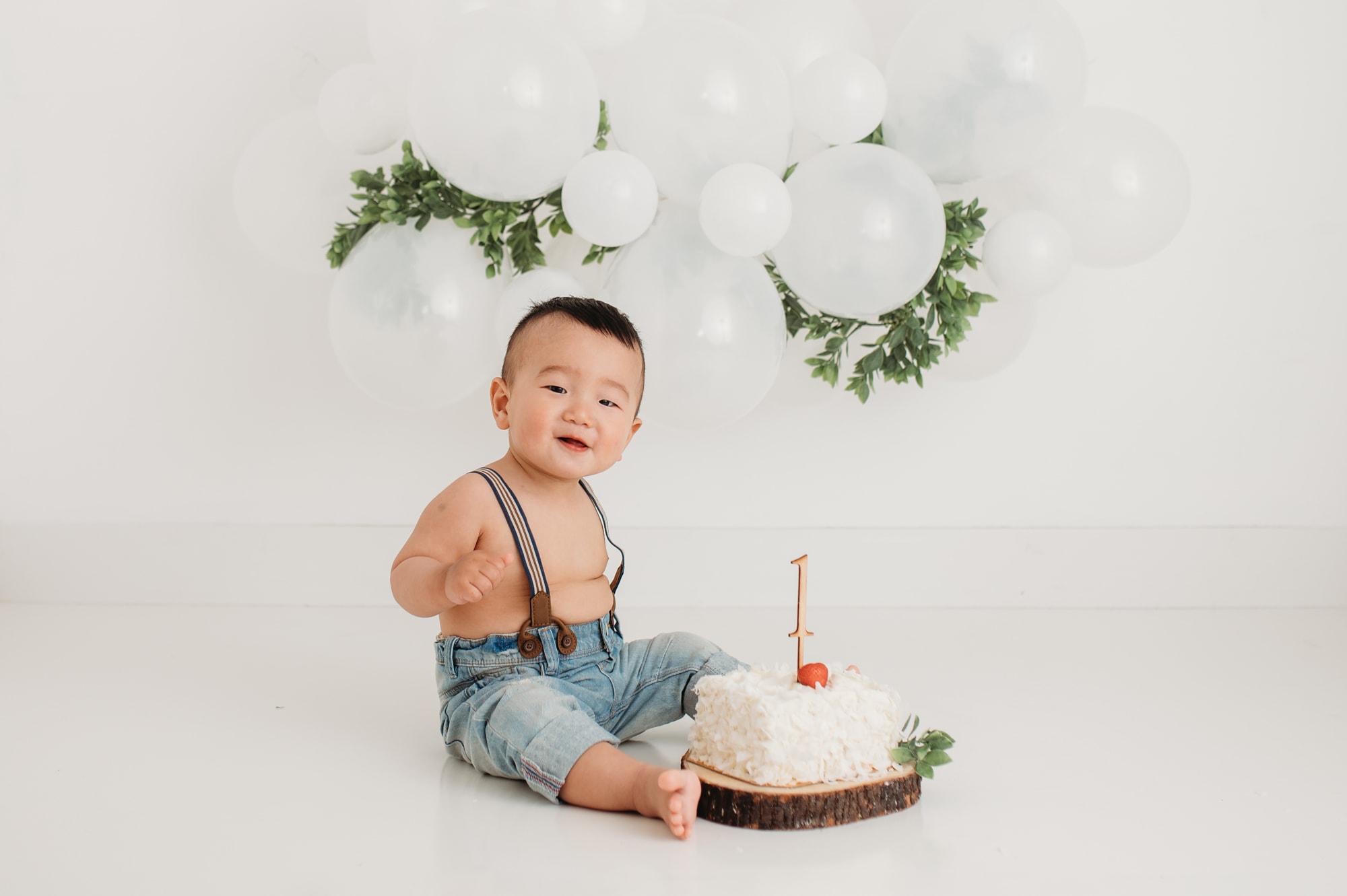Asian baby smiles at the camera during cake smash decorated with white ballon garland with green leaves.