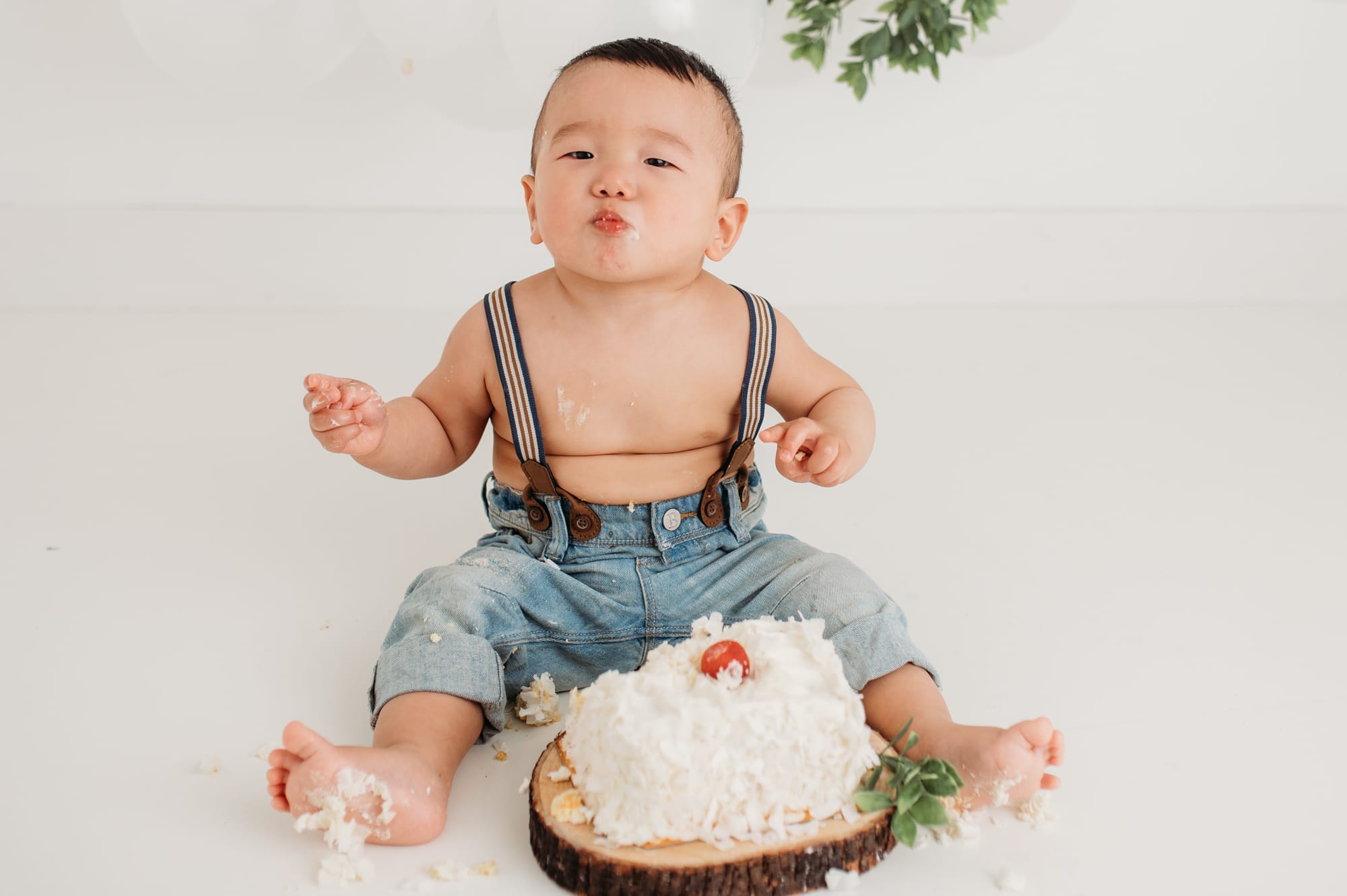 Baby blows kisses in blue denim and suspenders while eating cake in a Lower Mainland studio.