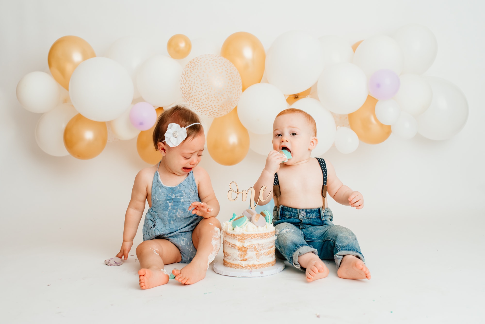 Funny cake smash for twins with white and gold balloon garland. Boy is eating cake while girl is crying.
