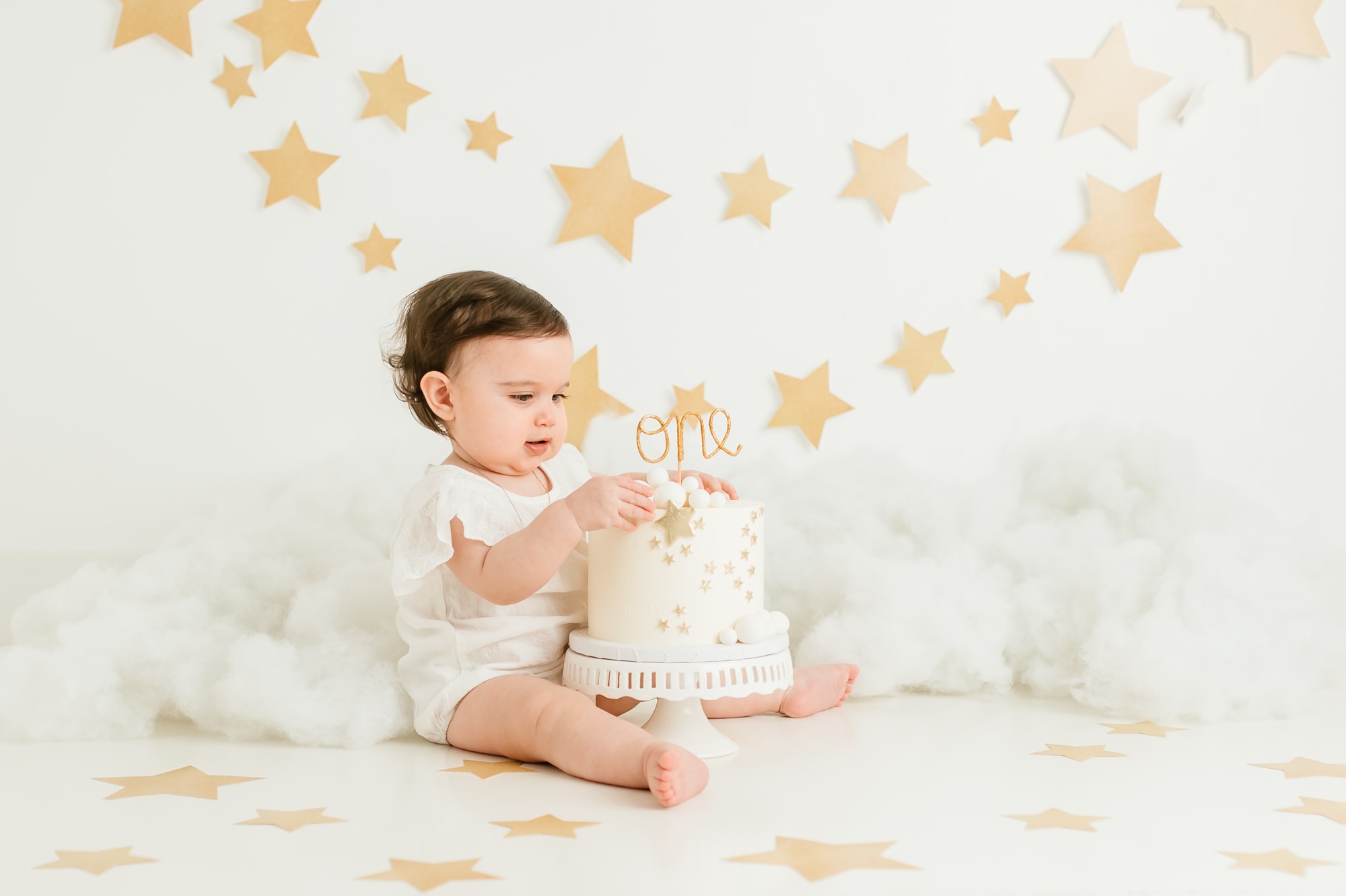 Girl reaching for a smash cake, with a cloud and stars background in white and gold.