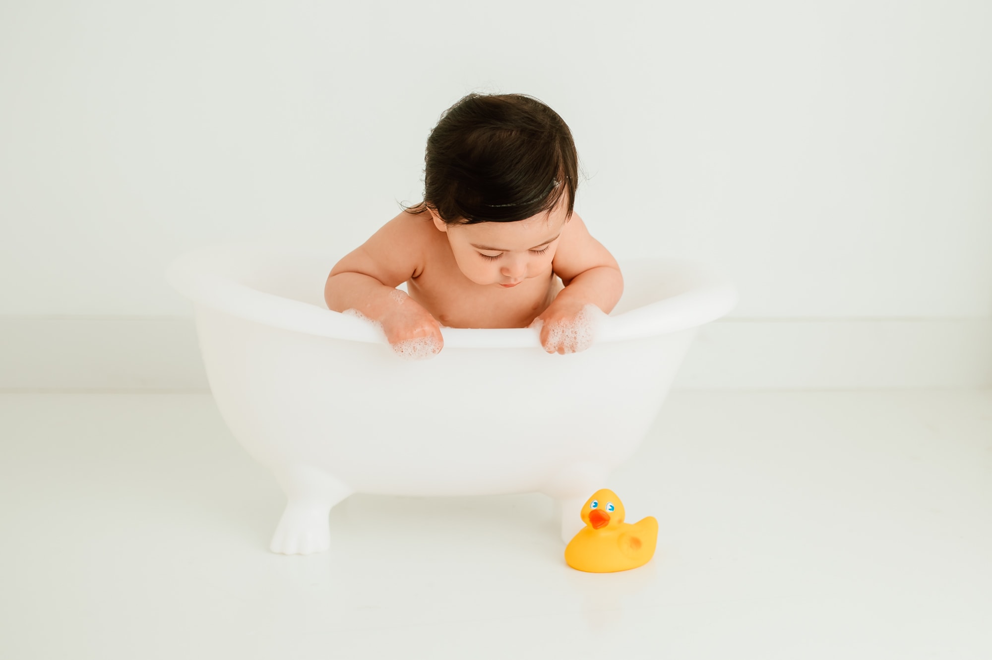 Baby taking a bath in a Denny's bathtub prop after cake smash and looking down at yellow rubber duck on the ground.