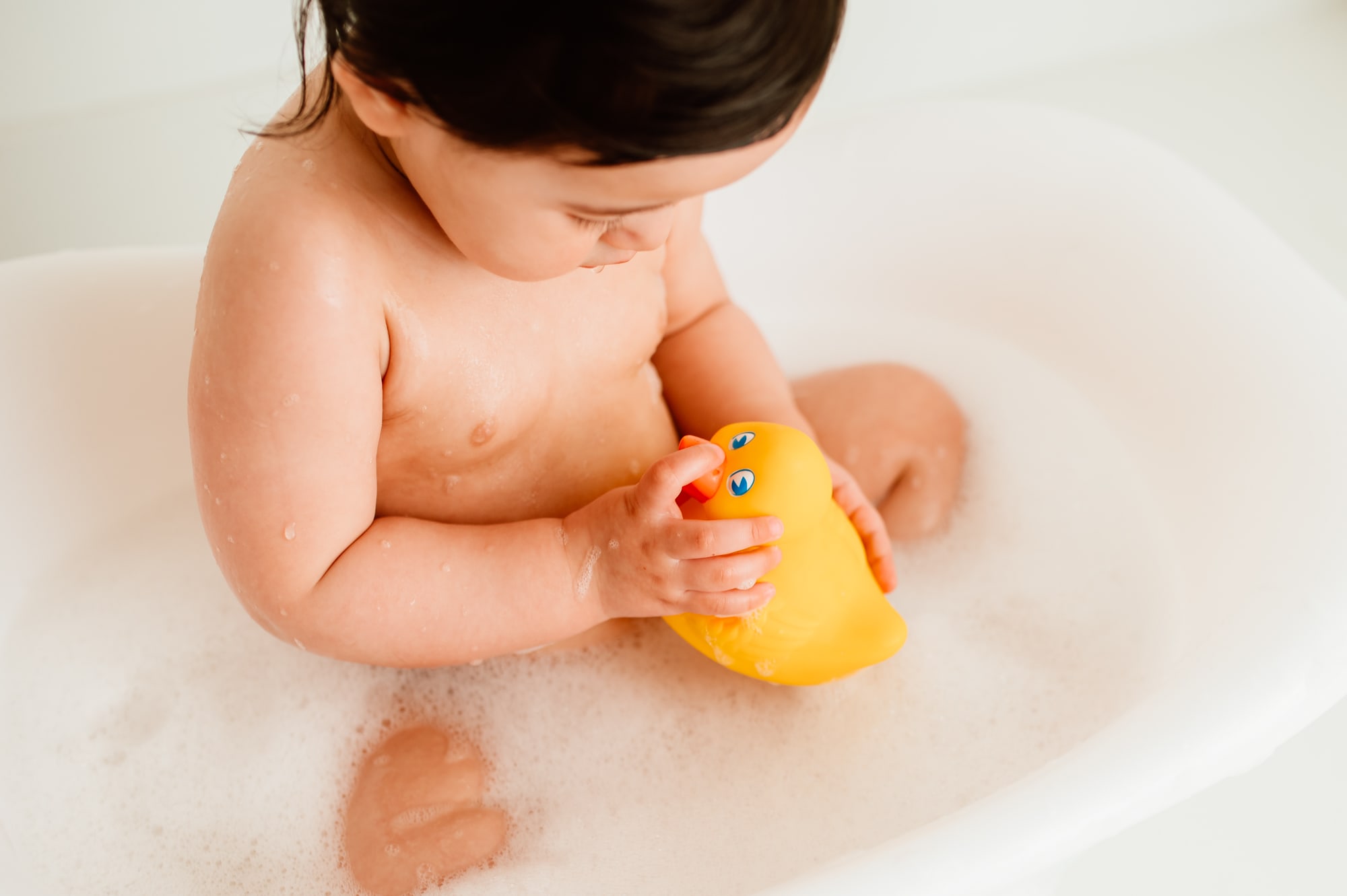 Child looks closely at rubber ducky during a bubble bath after their cake smash session.