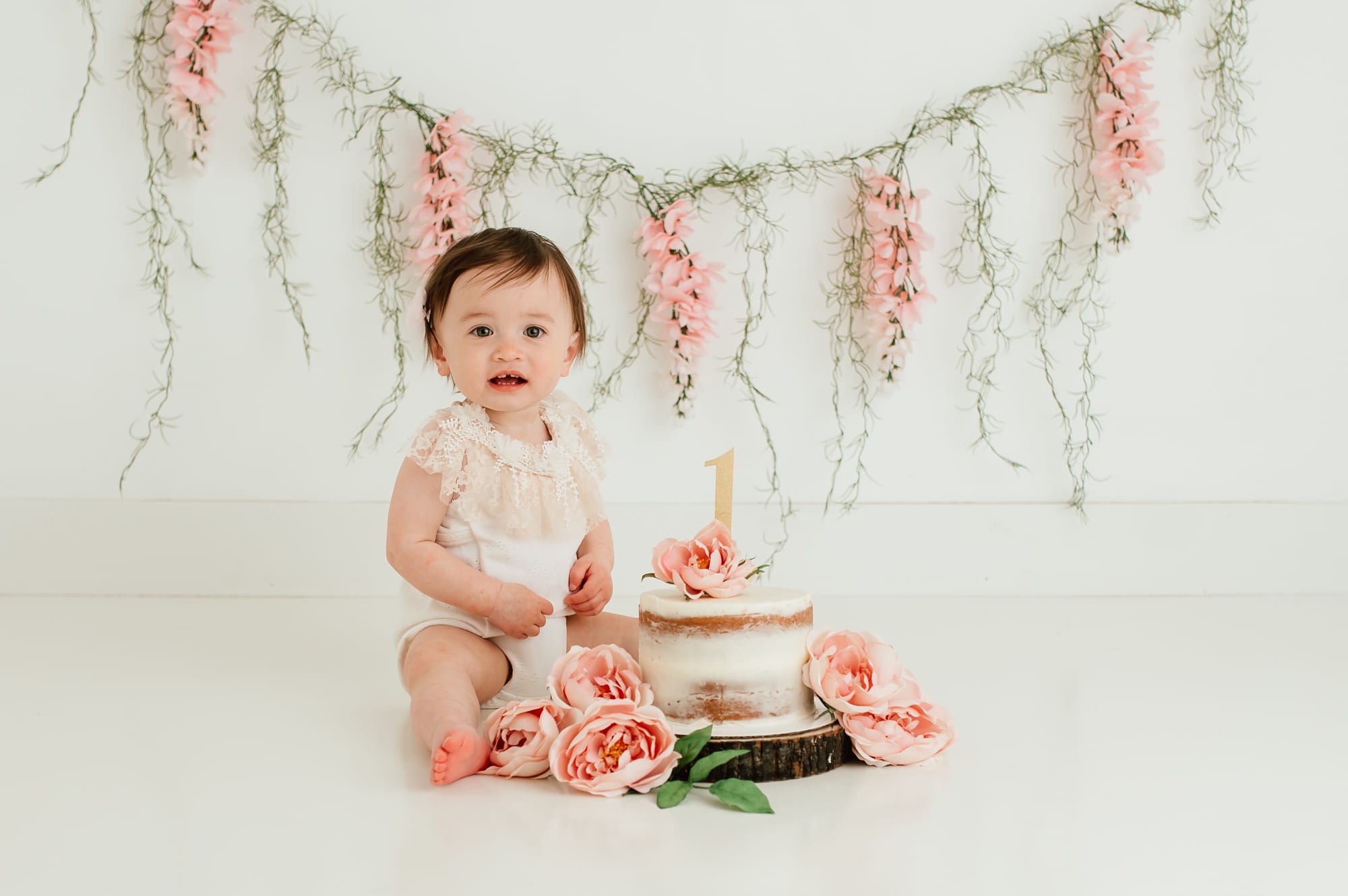 Floral and spanish moss backdrop behind one year old girl smiling during her Vancouver cake smash session.