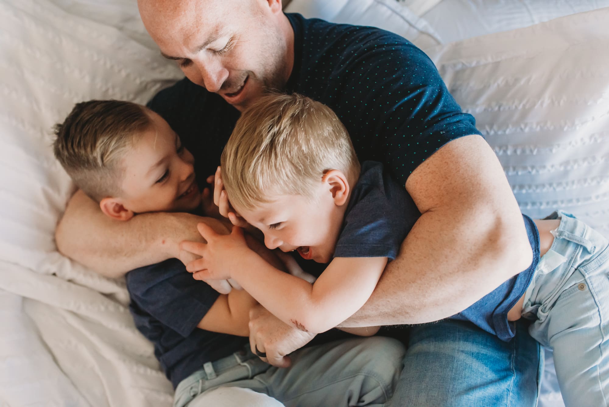 dad hugging boys while wrestling on bed shown by Vancouver Family Photographer