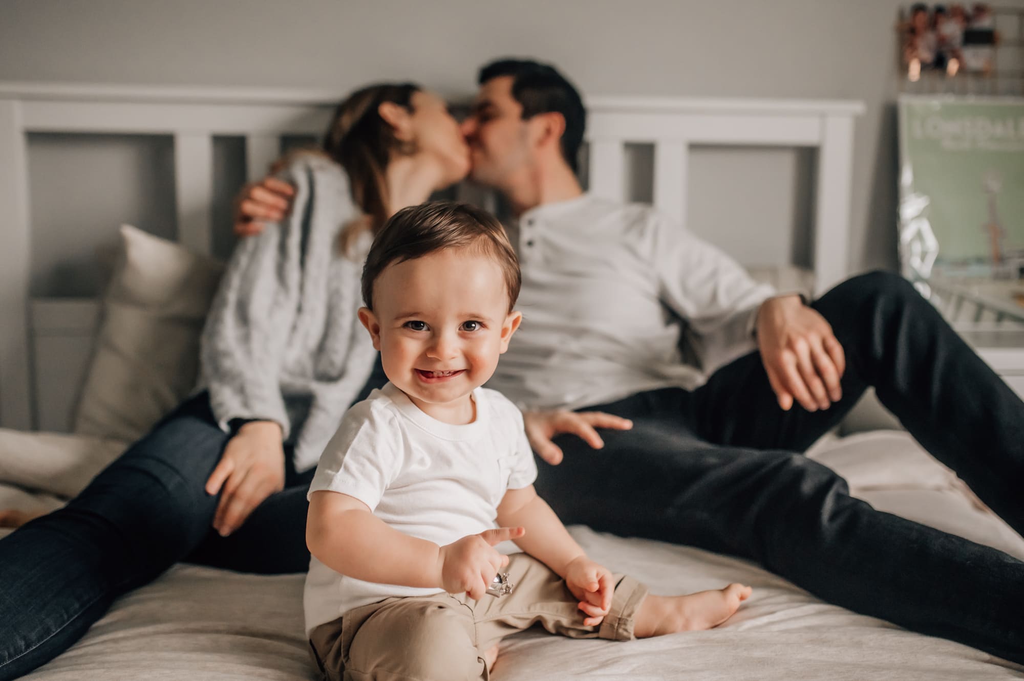 Inhome family photography at its best with laughing child sitting on the bed while parents are kissing in the background.