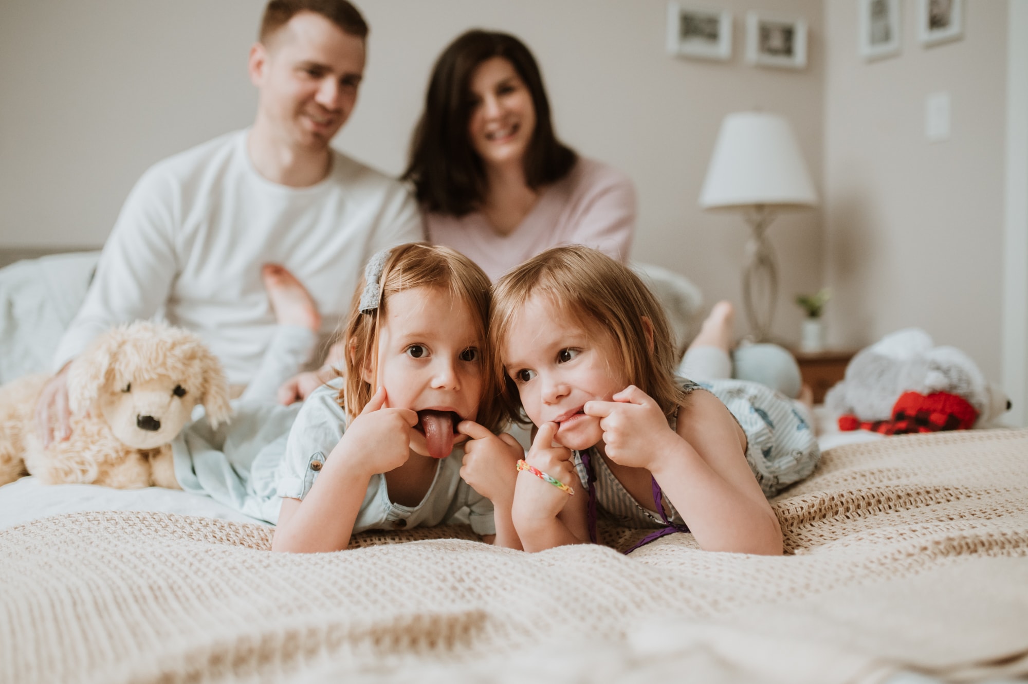 Twin girls pulling funny faces while lying on bed with parents looking on amused.