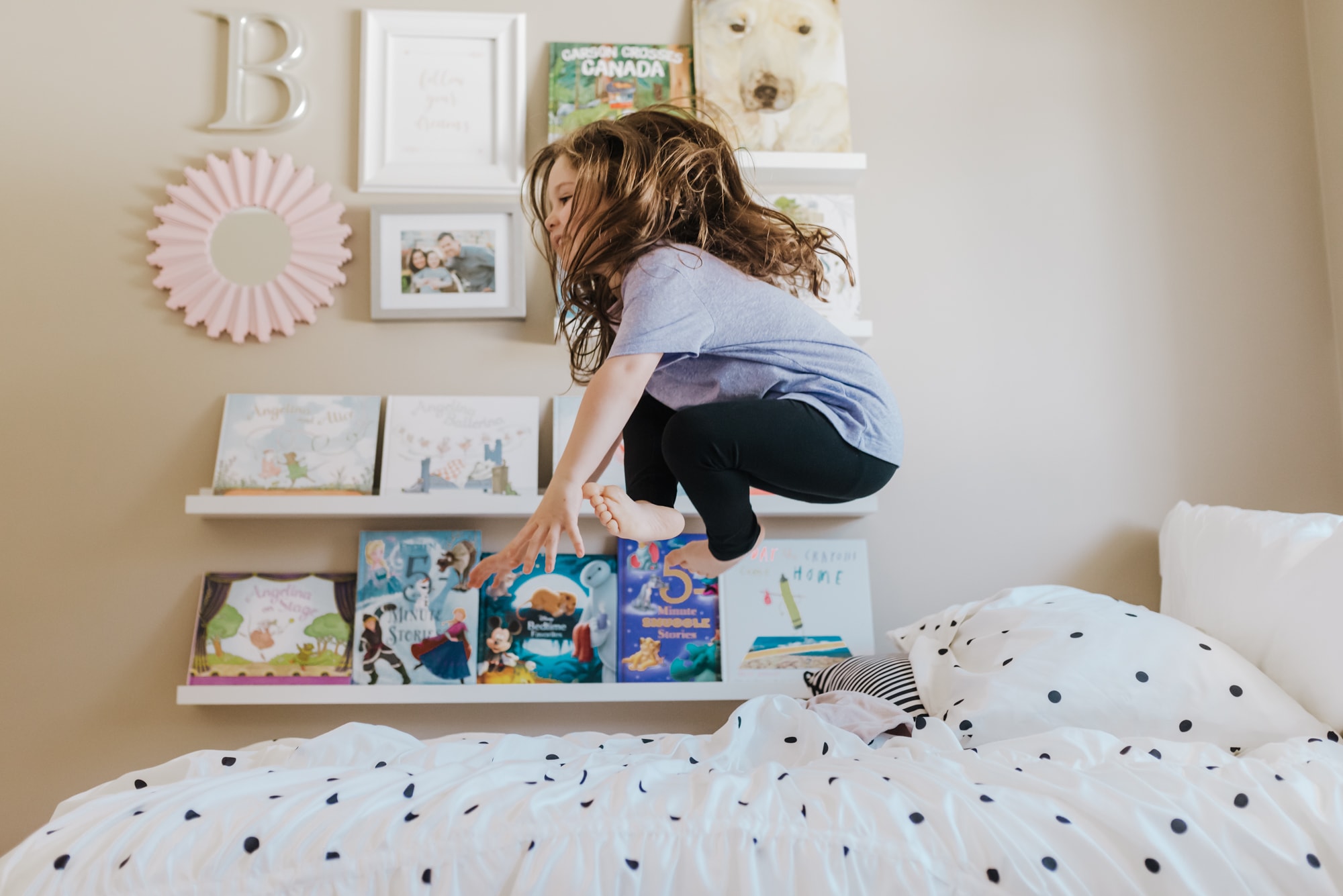 girl jumping on bed in bedroom with bookshelf displaying kids books during indoor family shoot