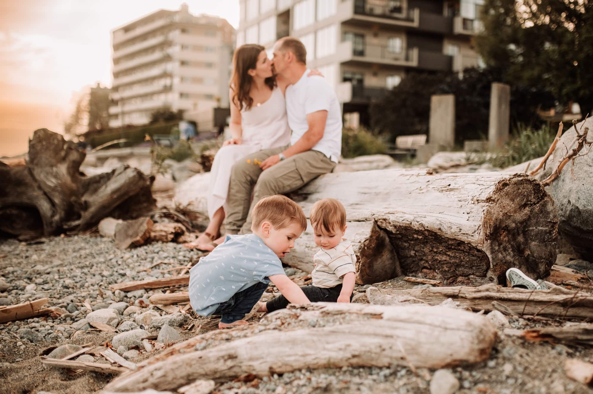 Family photoshoot in West Vancouver shows two small boys playing in the beach sand while their parents kiss in the background.